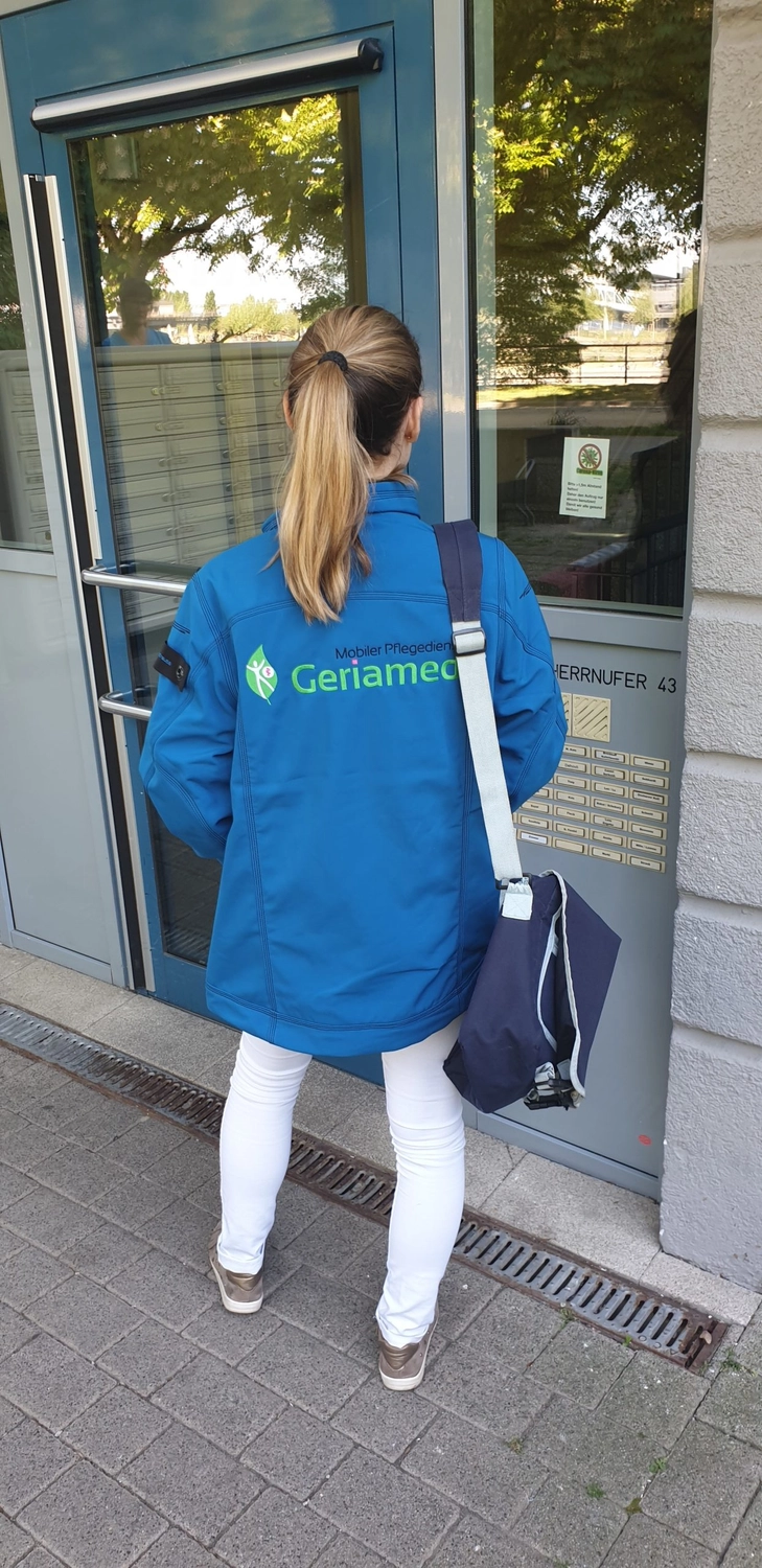 Geriamed GmbH – Ambulanter Pflegedienst – Eine Pflegekraft in einer blauen Geriamed-Jacke und einer weißen Hose steht am Eingang eines Gebäudes, eine dunkelblaue Tasche über der Schulter.