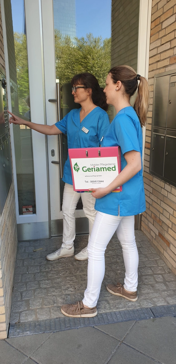 Geriamed GmbH - Ambulante Pflegedienst – Zwei Pflegekräfte in blauen OP-Kitteln stehen vor einem Gebäudeeingang; eine drückt auf die Türklingel, während die andere einen rosa Geriamed-Ordner in der Hand hält.