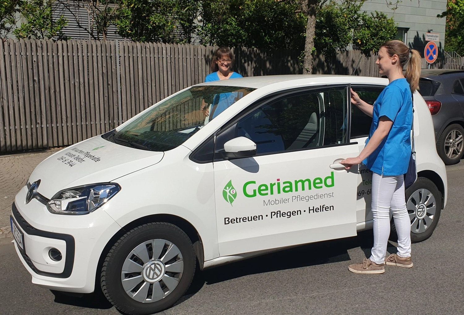 Geriamed GmbH – Ambulanter Pflegedienst – Zwei Pflegekräfte in blauen Kasacks stehen neben einem weißen Fahrzeug des ambulanten Pflegedienstes Geriamed, das in einer sonnigen Wohnstraße geparkt ist.