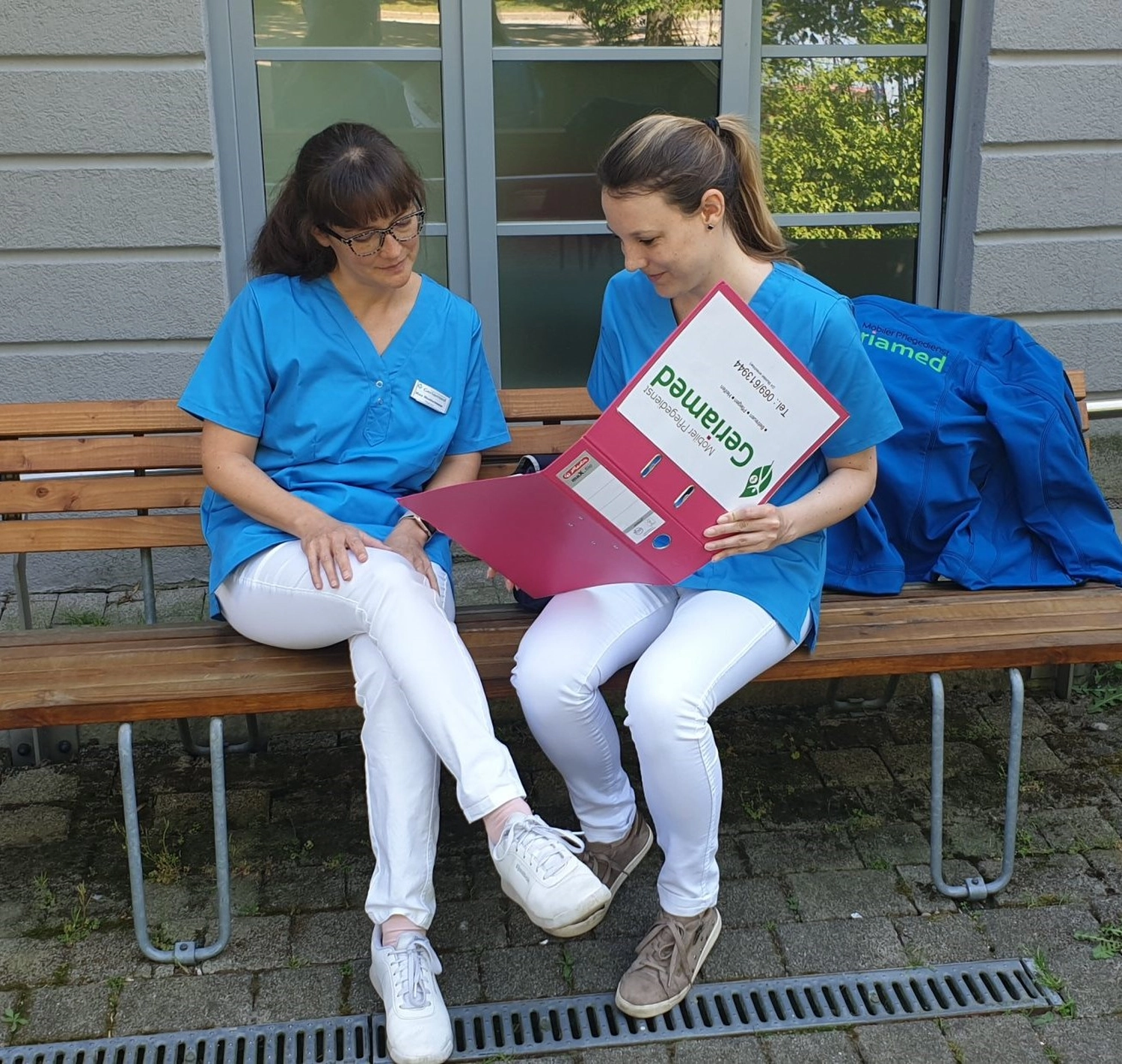 Geriamed GmbH – Ambulanter Pflegedienst – Zwei Pflegekräfte in blauen OP-Kitteln und weißen Hosen sitzen draußen auf einer Bank und sehen sich gemeinsam Unterlagen in einem rosa Geriamed-Ordner an.